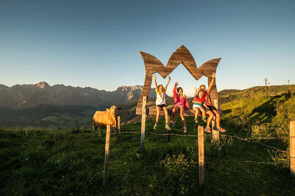 Kinder sitzen fröhlich auf einer Holzskulptur mit Bergblick, umgeben von Natur und Kühen.
